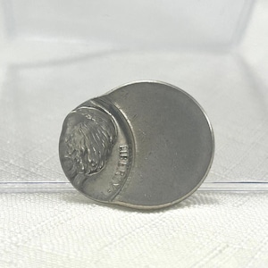 May include: A silver-colored coin-shaped object with a portrait of a man on the left side and the word "LIBERTY" visible. The coin has a circular shape with a slightly raised edge.