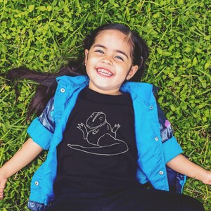 May include: A child wearing a black t-shirt with a white dinosaur graphic, a blue jacket, and black trousers, lying on green grass and smiling. The child is looking up at the camera.