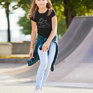 May include: A person wearing a black t-shirt with a white dinosaur graphic, light blue jeans, and a white baseball cap. They are standing on a pink skateboard with yellow wheels, with a green and blue plaid shirt tied around their waist.
