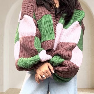 Può includere: Un cardigan lavorato all'uncinetto con un motivo a scacchi grandi in blocchi alternati di rosa chiaro, verde bosco e marrone scuro. Il capo presenta maniche lunghe e ampie con polsini a coste e un'apertura frontale con bottoni.