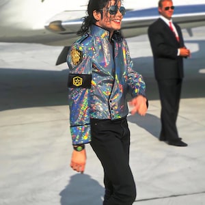 Puede incluir: Una chaqueta holográfica iridiscente con ribetes negros y un parche dorado en el hombro izquierdo. La chaqueta se combina con pantalones negros. Se usan gafas de sol. Una persona con traje está de pie en el fondo cerca de un avión.