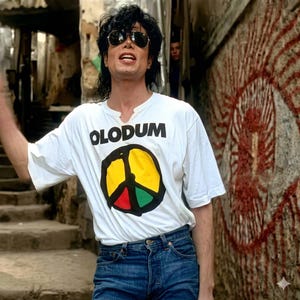 May include: A white t-shirt with the word "OLODUM" in black block letters and a peace sign in yellow, red, and green. The person is wearing sunglasses and blue jeans. The background shows a weathered wall with graffiti.