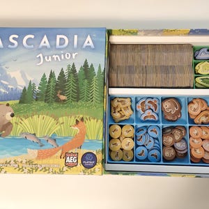 May include: Open board game box titled "Cascadia Junior." The box art features a bear, fox, and fish in a natural landscape. Inside, the game includes cards and various animal-shaped tokens in blue and green compartments.