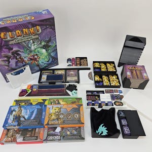 May include: Clank! Catacombs board game box and components. The box is purple and features fantasy artwork. Includes cards, tokens, and various game pieces in black trays and containers. A white box and black bag are also included.