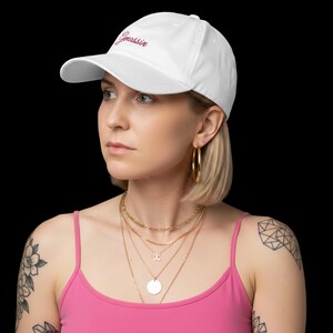 Könnte beinhalten: Eine weiße Baseballkappe mit dem Wort "Gnossin" in kursiver, magenta Schrift gestickt. Die Kappe hat einen gebogenen Schirm und ein klassisches Sechs-Panel-Design. Der Hintergrund ist schwarz.