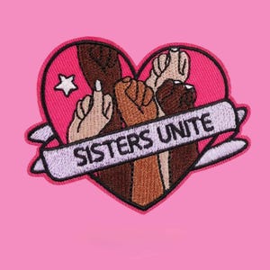 Könnte beinhalten: Ein herzförmiger Stickaufnäher mit rosa Hintergrund, einem weißen Stern und mehreren erhobenen Fäusten in verschiedenen Hauttönen. Ein hellviolettes Banner in der Mitte zeigt den schwarzen Text SISTERS UNITE in einer fetten, serifenlosen Schrift.