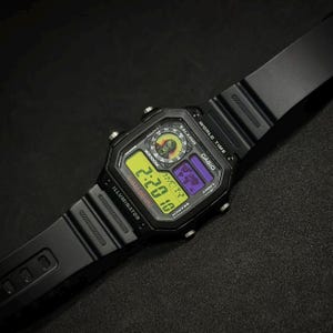 Könnte beinhalten: Schwarze digitale Casio-Uhr mit rechteckigem Zifferblatt und schwarzem Armband. Das Zifferblatt zeigt Uhrzeit, Datum und Weltzeitfunktionen an. Das Zifferblatt hat eine gelbe, lila und grüne Anzeige. Die Uhr trägt die Aufschrift "CASIO" und "ILLUMINATOR".