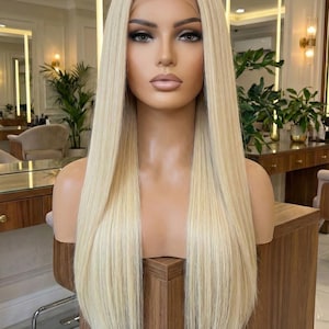May include: A blonde wig with long, straight hair is displayed on a mannequin head. The wig has a natural-looking hairline and a light blonde color. The mannequin is positioned on a wooden surface, and the background shows a salon setting.
