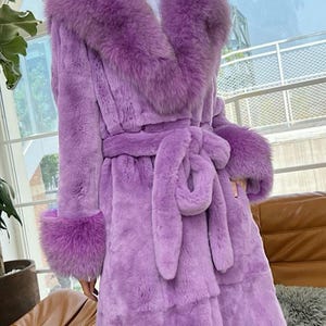 May include: A long, lavender faux fur coat with a large collar and cuffs. The coat features a matching belt tied at the waist. The coat is a luxurious outerwear garment.