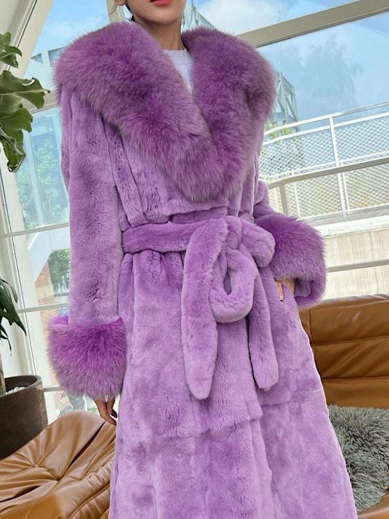 May include: A long, lavender faux fur coat with a large collar and cuffs. The coat features a matching belt tied at the waist. The coat is a luxurious outerwear garment.