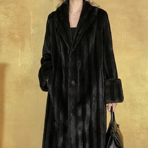 May include: A long, black fur coat with a classic collar and cuffs. The coat is floor-length and has a vertical striped pattern. A black handbag is held in the hand. The background is a solid gold color.