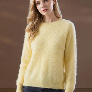 May include: A pale yellow crew neck sweater with long sleeves. The sweater has a soft, fuzzy texture and is paired with dark blue jeans. The model has long blonde hair.