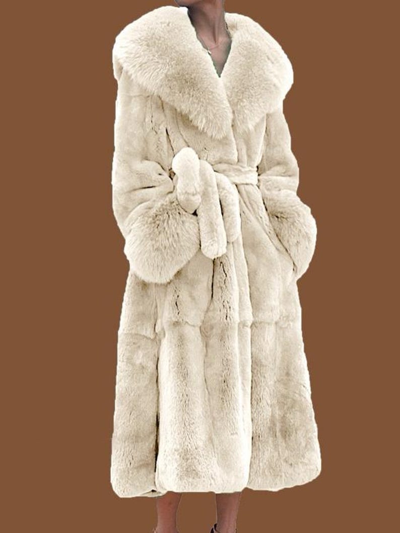 May include: Cream-coloured faux fur coat featuring a large collar, long sleeves, and a matching belt. The coat has a knee-length hemline and a soft, textured appearance. The coat is designed for warmth and style.
