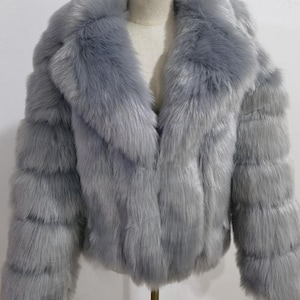 May include: A light gray faux fur jacket with a wide collar and long sleeves. The jacket is displayed on a mannequin and has a soft, textured appearance. The jacket is a stylish outerwear option.