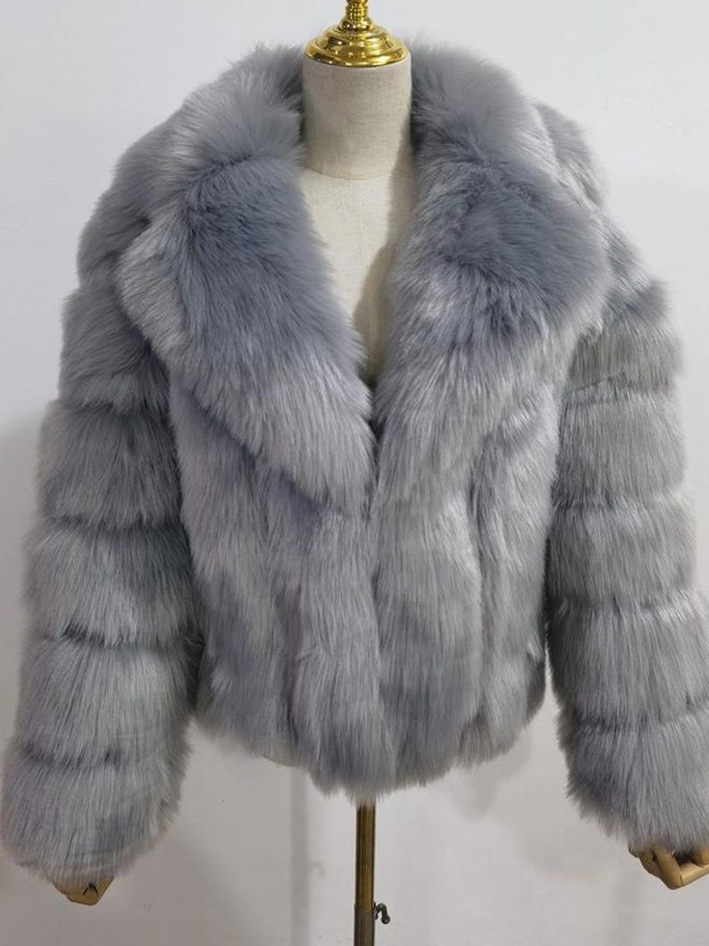 May include: A light gray faux fur jacket with a wide collar and long sleeves. The jacket is displayed on a mannequin and has a soft, textured appearance. The jacket is a stylish outerwear option.