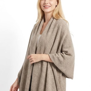 Puede incluir: Un suéter tipo capa beige con cuello drapeado y mangas anchas. La prenda de punto suave se lleva sobre una camiseta blanca. La modelo sonríe, con el pelo largo y rubio, sobre un fondo blanco.