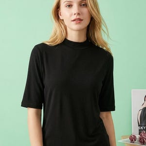 Könnte beinhalten: Ein schwarzes Oberteil mit kurzem Arm und Stehkragen. Das Oberteil ist aus einem weichen, glatten Stoff gefertigt. Das Model trägt das Oberteil. Der Hintergrund ist hellgrün.