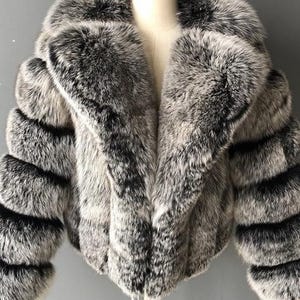 May include: A faux fur jacket in shades of gray and black, displayed on a mannequin. The jacket features a wide collar and horizontal stripes on the sleeves. The overall style is a cropped, short length.