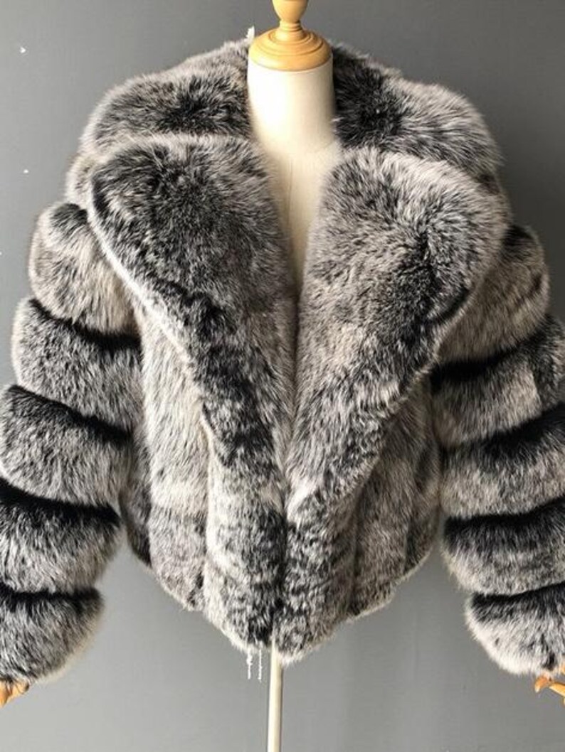 May include: A faux fur jacket in shades of gray and black, displayed on a mannequin. The jacket features a wide collar and horizontal stripes on the sleeves. The overall style is a cropped, short length.