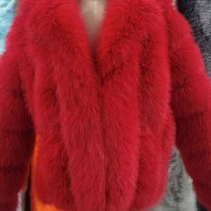 May include: A vibrant red faux fur jacket with a wide collar and long sleeves. The jacket is displayed on a mannequin, showcasing its plush texture and bold color. The jacket's design features a V-neckline and a cropped length.