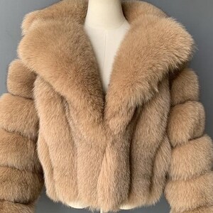 May include: A tan-colored faux fur jacket with a wide collar and three-quarter sleeves. The jacket is displayed on a mannequin and has a soft, textured appearance. The sleeves are designed with a layered, bubble-like effect.