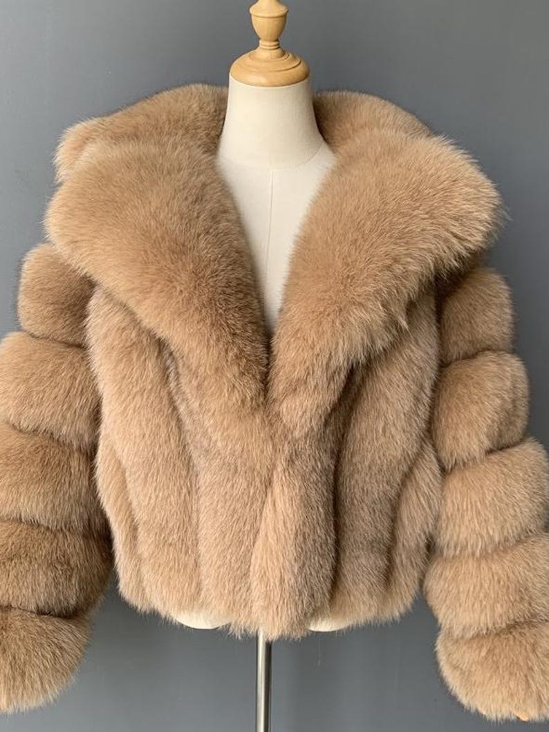 May include: A tan-colored faux fur jacket with a wide collar and three-quarter sleeves. The jacket is displayed on a mannequin and has a soft, textured appearance. The sleeves are designed with a layered, bubble-like effect.