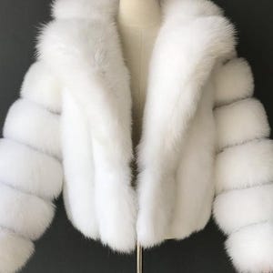 May include: A white faux fur jacket with a large collar and long sleeves. The jacket is displayed on a mannequin. The sleeves have a unique design with multiple horizontal sections. The jacket is a winter outerwear item.