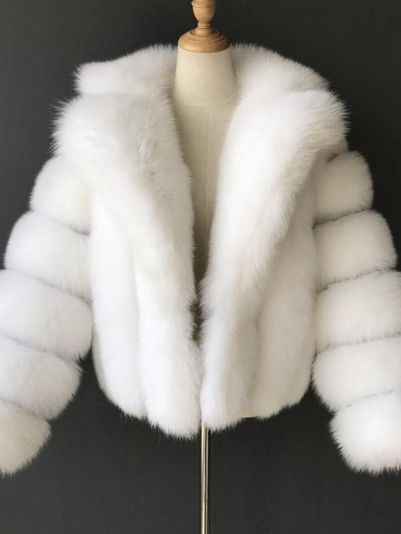 May include: A white faux fur jacket with a large collar and long sleeves. The jacket is displayed on a mannequin. The sleeves have a unique design with multiple horizontal sections. The jacket is a winter outerwear item.