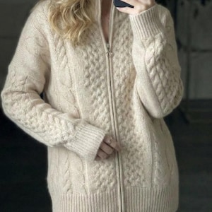 May include: Cream-colored cable knit zip-up sweater. The sweater features a high collar and ribbed cuffs and hem. The front has a central zipper and a textured cable knit pattern. The sleeves also have a cable knit design. The sweater is a neutral color.