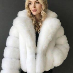 May include: A white faux fur jacket with a wide collar and long sleeves. The jacket has a fluffy, textured appearance and a cropped length. The model is wearing a black top underneath. The jacket is a winter outerwear item.