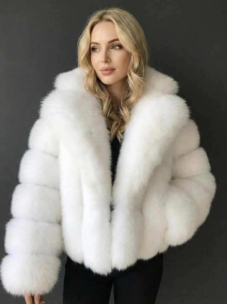 May include: A white faux fur jacket with a wide collar and long sleeves. The jacket has a fluffy, textured appearance and a cropped length. The model is wearing a black top underneath. The jacket is a winter outerwear item.