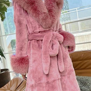 May include: A long, pink faux fur coat with a matching fur collar, cuffs, and belt. The coat features a wrap-around design with a tie belt at the waist. The coat is a solid pink colour and appears to be made of a soft, plush material.