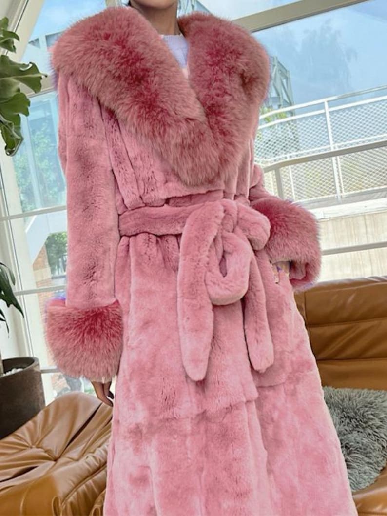 May include: A long, pink faux fur coat with a matching fur collar, cuffs, and belt. The coat features a wrap-around design with a tie belt at the waist. The coat is a solid pink colour and appears to be made of a soft, plush material.