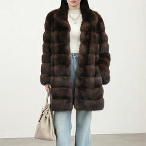 May include: A brown fur coat with a wide collar and horizontal panel design. The coat is worn over a white top and blue jeans. A light-colored handbag is held in the left hand.