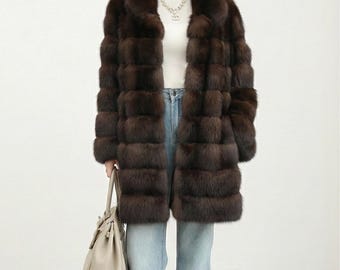 May include: A brown fur coat with a wide collar and horizontal panel design. The coat is worn over a white top and blue jeans. A light-colored handbag is held in the left hand.