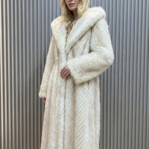 May include: A long, cream-coloured faux fur coat with a large hood. The coat has a textured, chevron-like pattern and a relaxed fit. The model is wearing the coat, a stylish outerwear piece.