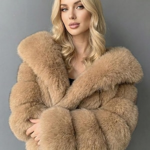 May include: A tan-colored fur coat with a wide collar and sleeves. The coat has a plush, textured appearance and is worn over a black top. The background is a solid gray.