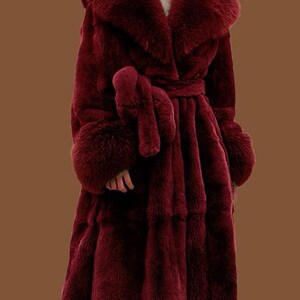 May include: A long, burgundy faux fur coat with a wide collar and matching cuffs. The coat features a tie belt at the waist and a full, flowing skirt. The plush texture and rich colour create a luxurious look.