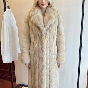 May include: A long, beige faux fur coat with a wide collar. The coat is floor-length and has a vertical ribbed texture. A white shirt hangs on a rack in the background.