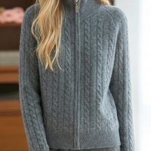 May include: A gray zip-up sweater with a high collar. The sweater has a cable knit design and a gold-colored zipper. The sleeves and hem have ribbed detailing. The sweater is made of a soft, textured material.