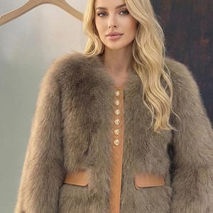 May include: A brown fur coat with a round collar and long sleeves. The coat features a tan leather panel down the front with gold buttons and two front pockets. The coat is a neutral color and appears soft and luxurious.