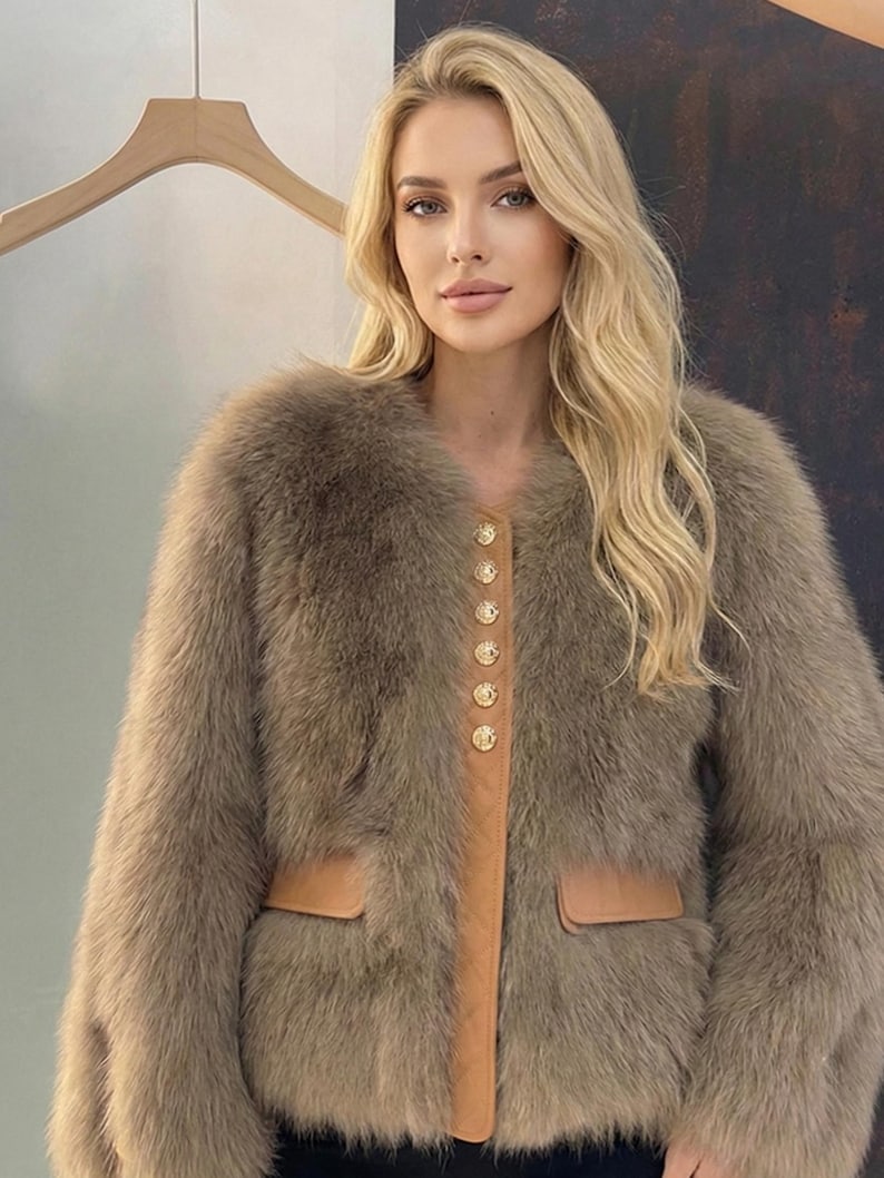May include: A brown fur coat with a round collar and long sleeves. The coat features a tan leather panel down the front with gold buttons and two front pockets. The coat is a neutral color and appears soft and luxurious.