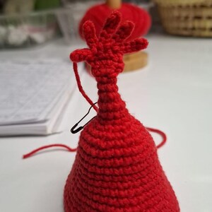 May include: A handmade, red crocheted figure with a bell-shaped base and a hand-like top. A black safety pin and red yarn are attached. The figure is on a white surface, with a wooden spool and red yarn in the background.