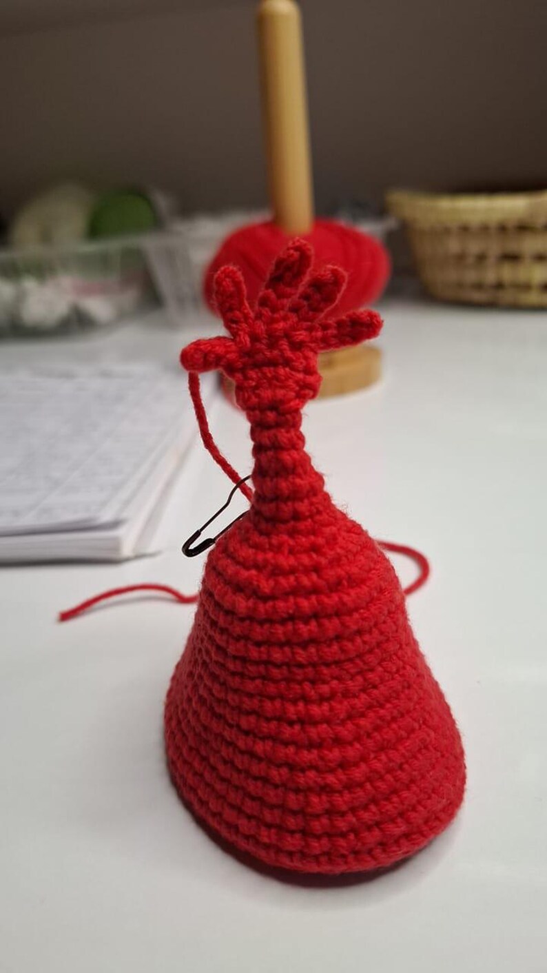 May include: A handmade, red crocheted figure with a bell-shaped base and a hand-like top. A black safety pin and red yarn are attached. The figure is on a white surface, with a wooden spool and red yarn in the background.