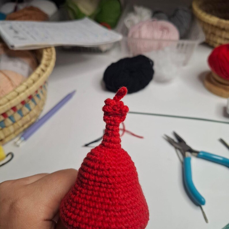 May include: A close-up of a handmade red crochet item, possibly a doll or ornament, with a bulbous body and a slender neck. The top features a detailed, knotted design. Crafting tools and yarn are visible in the background.