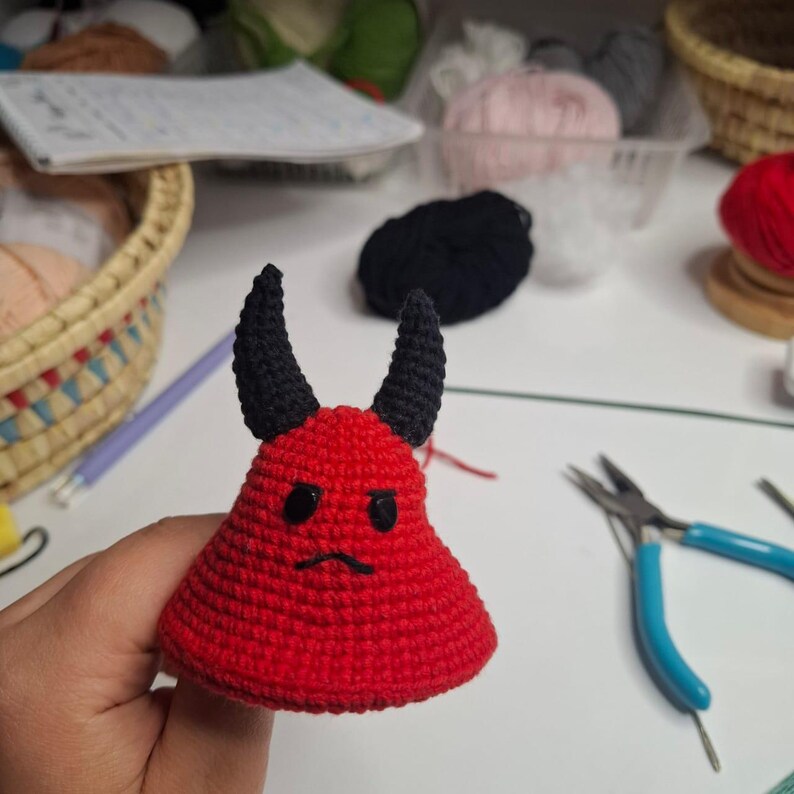 May include: A handmade red crochet devil with black horns and a small, sad face. The devil is a small, cone-shaped figure, likely a toy or decorative item. The background includes craft supplies.