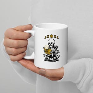 Può includere: Tazza in ceramica bianca con un disegno di uno scheletro che legge un libro, con dettagli dorati e fasi lunari. La tazza è tenuta da una persona.
