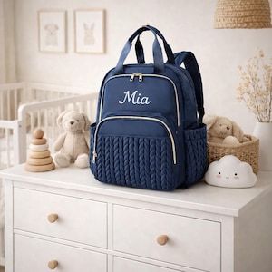 May include: Navy blue baby changing backpack with gold zips and trim. The bag has a braided texture on the front pocket and the name "Mia" embroidered in white. Includes top handles and adjustable shoulder straps.