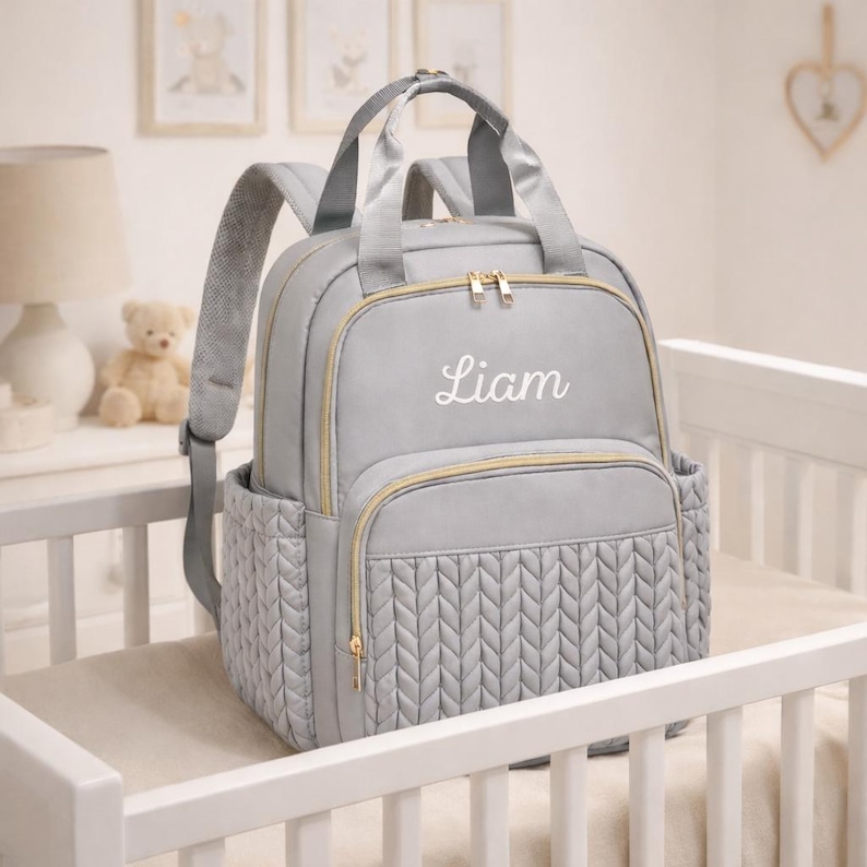 Gray personalized diaper backpack with embroidered name liam displayed on baby crib