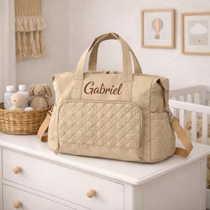 Pode incluir: Bolsa de fraldas bege com o nome "Gabriel" bordado na frente. A bolsa tem um bolso frontal acolchoado, alças superiores e uma alça de ombro. Uma cesta de vime com artigos para bebês e um bicho de pelúcia estão no fundo.